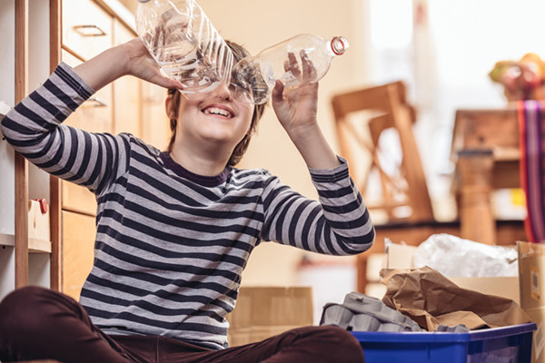 Caucasian boy sitting on the kitchen floor, sorting out the recycling in to blue box. He is holding two plastic bottles on his face, pretending to look thru binoculars. Sunny day interior.