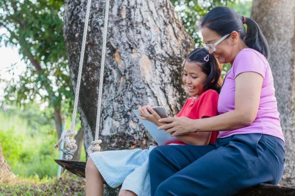 A woman and child reading a book next to a tree on an outdoor swing.