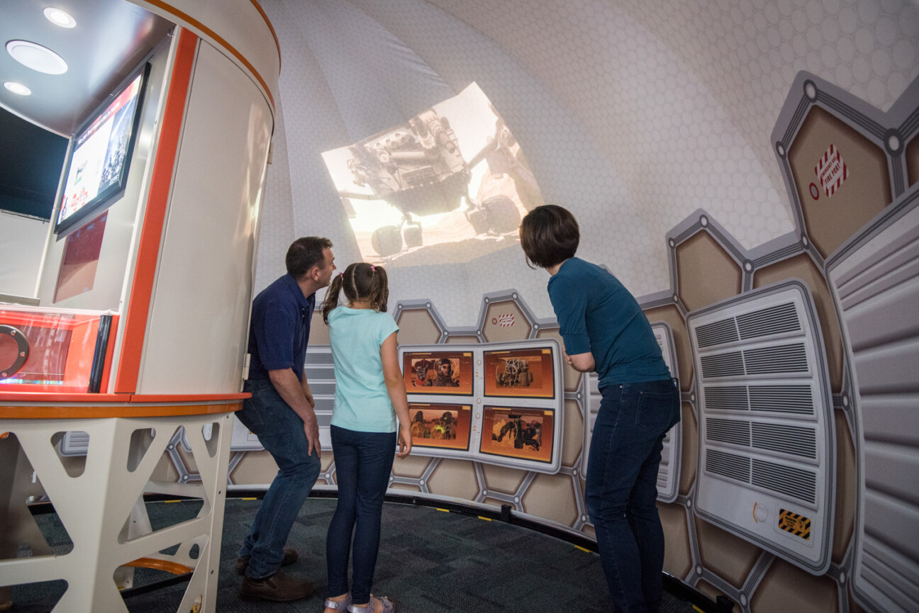 Two adults and a child watching a Mars rover on a projector screen