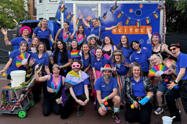 Scitech staff, friends and family in blue Scitech shirts, glitter and pride flags smile happily in front of a Scitech truck.